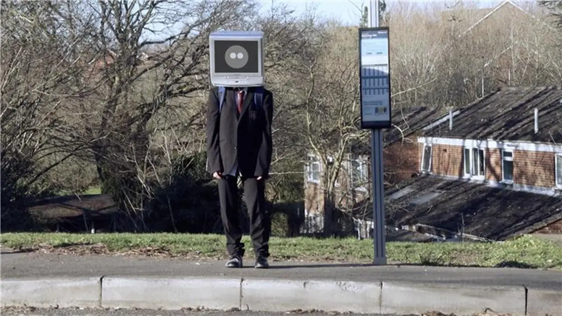 Boy standing at a bus stop with a telly monitor on his head.